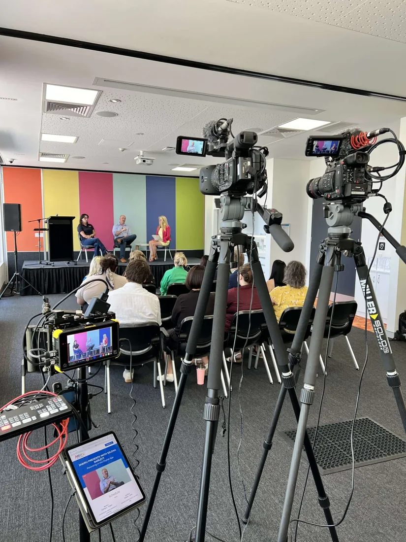 Three cameras on tripods covering a three-person panel discussion in front of a seated audience